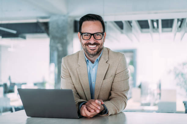 istockphoto-2153072054-612×612 Shot of a businessman sitting on desk in modern office and working on laptop. Elegant handsome businessman sitting in his office and using laptop.