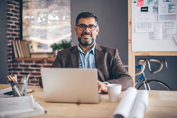 istockphoto-499335798-612×612 Mature business man working at modern office space. Sitting at the table and working on laptop. Looking confidently at camera. Brick wall and bicycle in background.